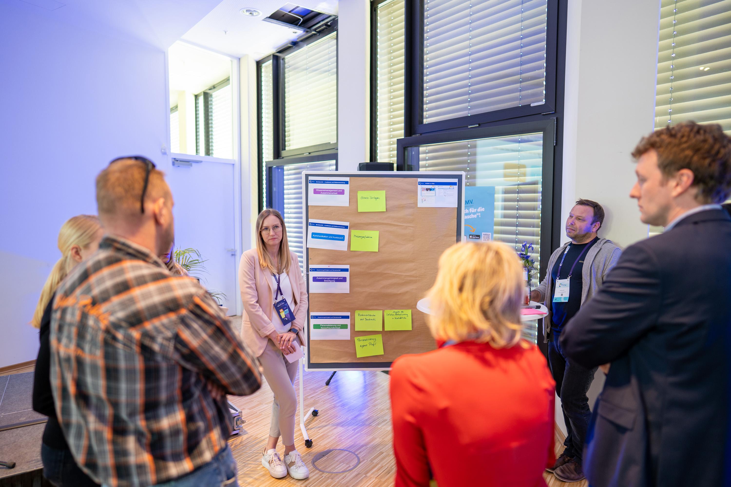 Gruppe von Menschen, die um ein Flipchart mit Notizen stehen. Eine Frau in heller Kleidung präsentiert.