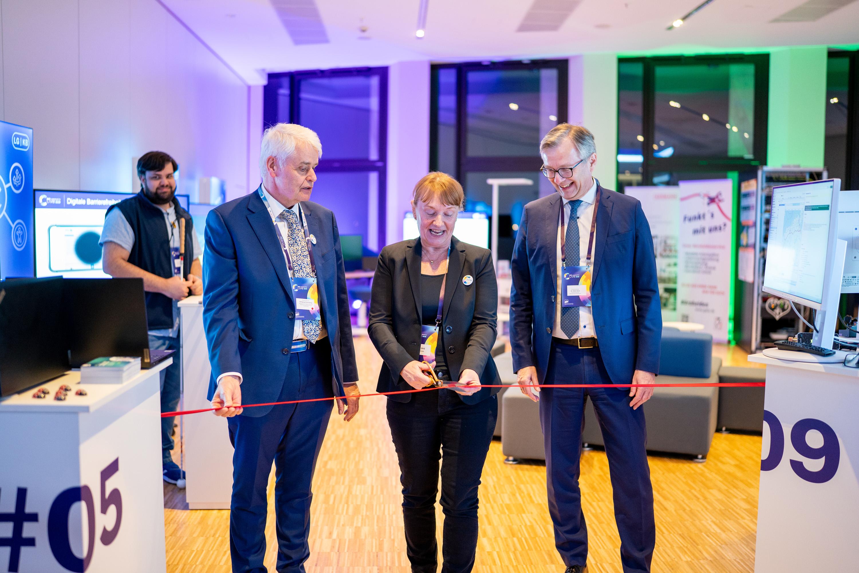 Finanzminister Dr. Heiko Geue, Staatssekretär Wolfgang Schmülling und Uta Knöchel schneiden ein rotes Band durch, um dieVeranstaltung zu eröffnen. Im Hintergrund sind Stände und Displays zu sehen.