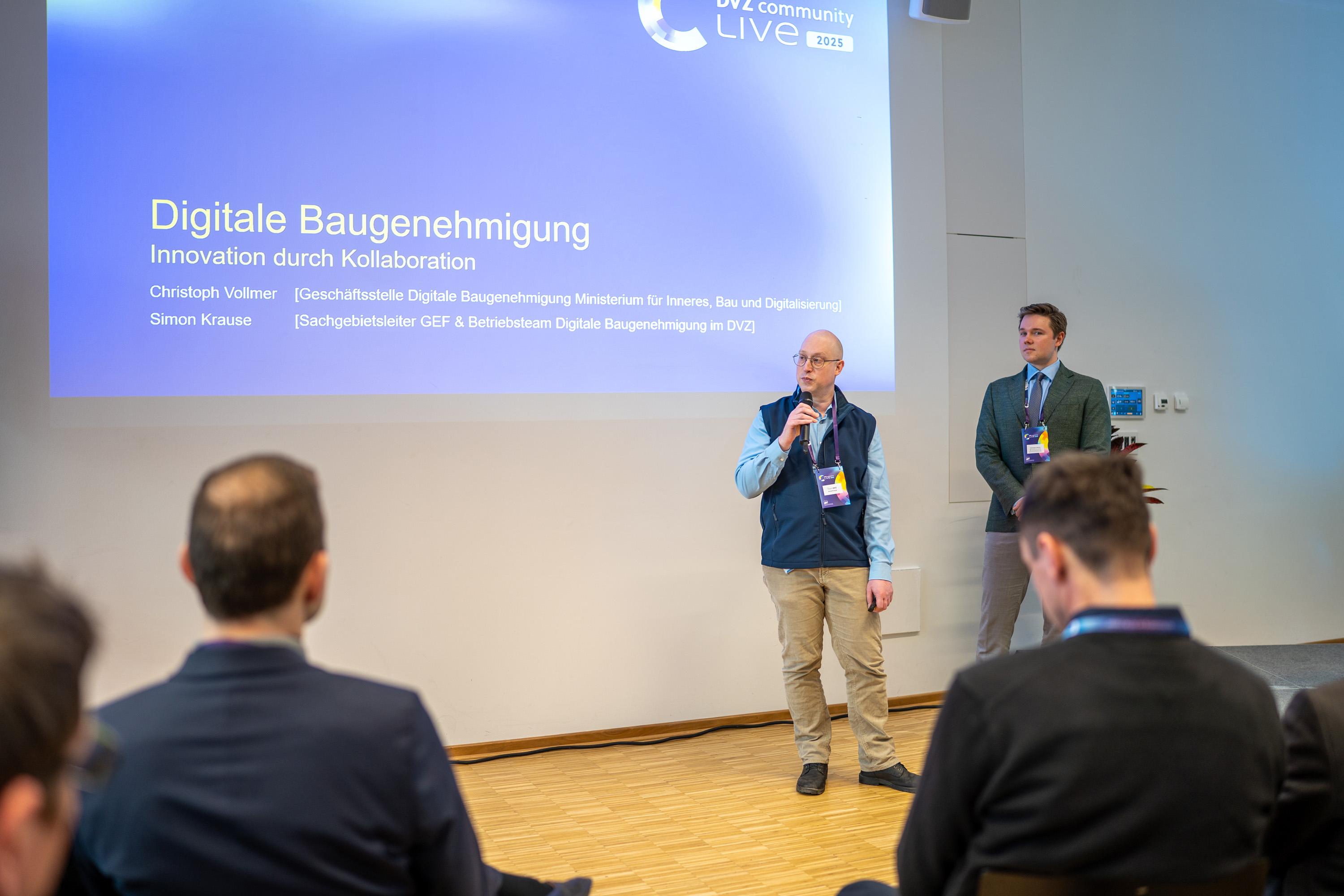 Zwei Männer präsentieren auf einer Bühne im Raum Mecklenburg. Der erste Mann spricht ins Mikrofon, der zweite steht daneben. Im Hintergrund ist eine Präsentationsfolie mit dem Titel "Digitale Baugenehmigung" zu sehen.