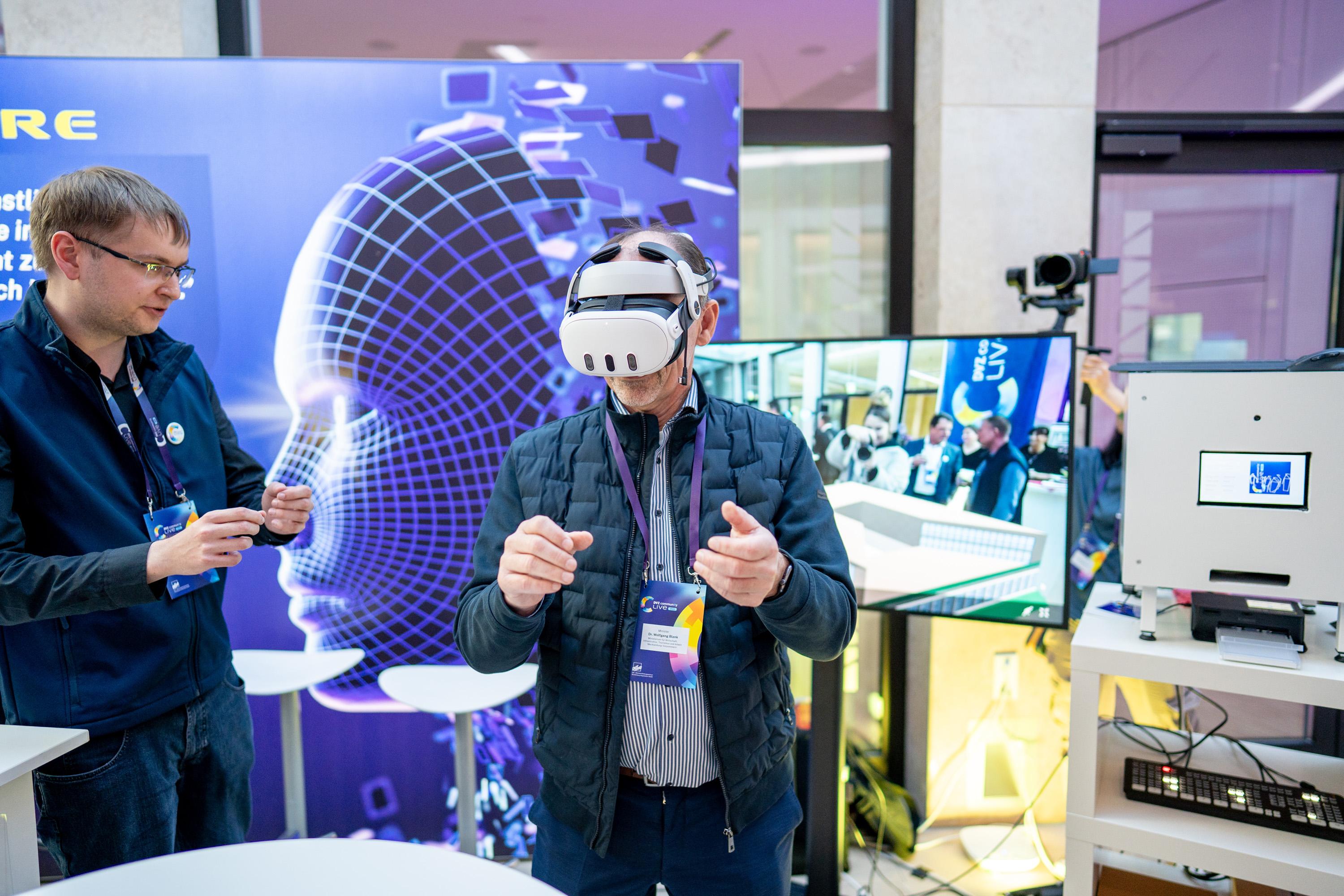 Wirtschaftsminister Wolfgang Blank trägt eine VR-Brille und interagiert mit einer virtuellen Umgebung. Im Hintergrund steht ein Bildschirm mit einer Präsentation.